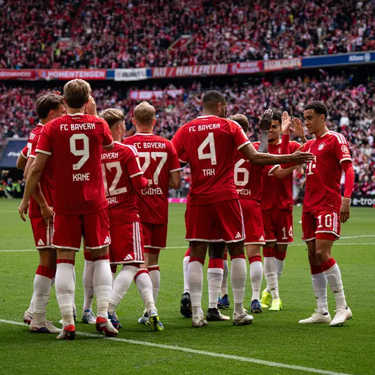 A group of FC Bayern Munich players in red and white uniforms is gathered on the field, celebrating and interacting with one another. The stadium is filled with fans in the background, creating a vibrant atmosphere.