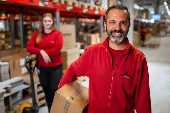 A man in a red jacket is smiling while carrying a cardboard box. Behind him, a woman in a red shirt is standing next to a pallet jack, with shelves of boxes visible in the warehouse setting.