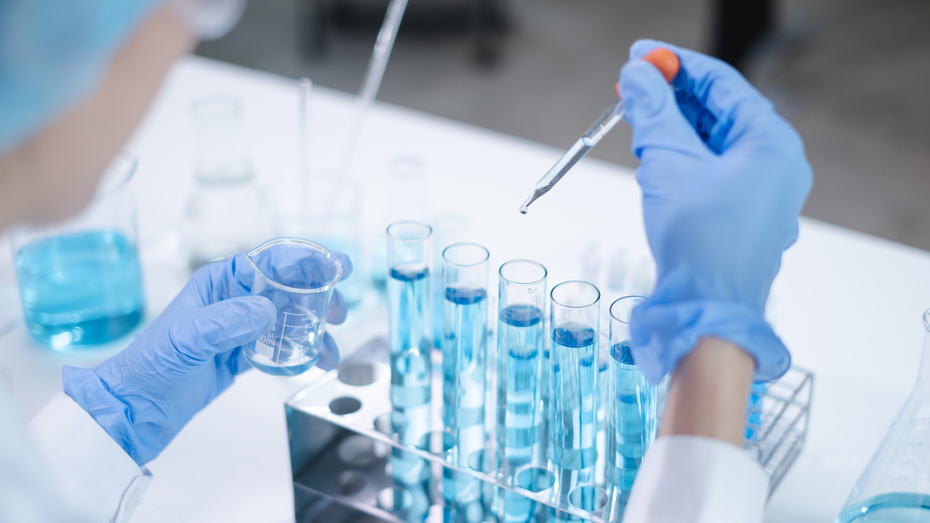 A scientist in a lab coat and blue gloves is holding a small beaker in one hand and using a dropper to transfer a liquid into test tubes filled with blue liquid. The lab environment features various glassware and equipment.
