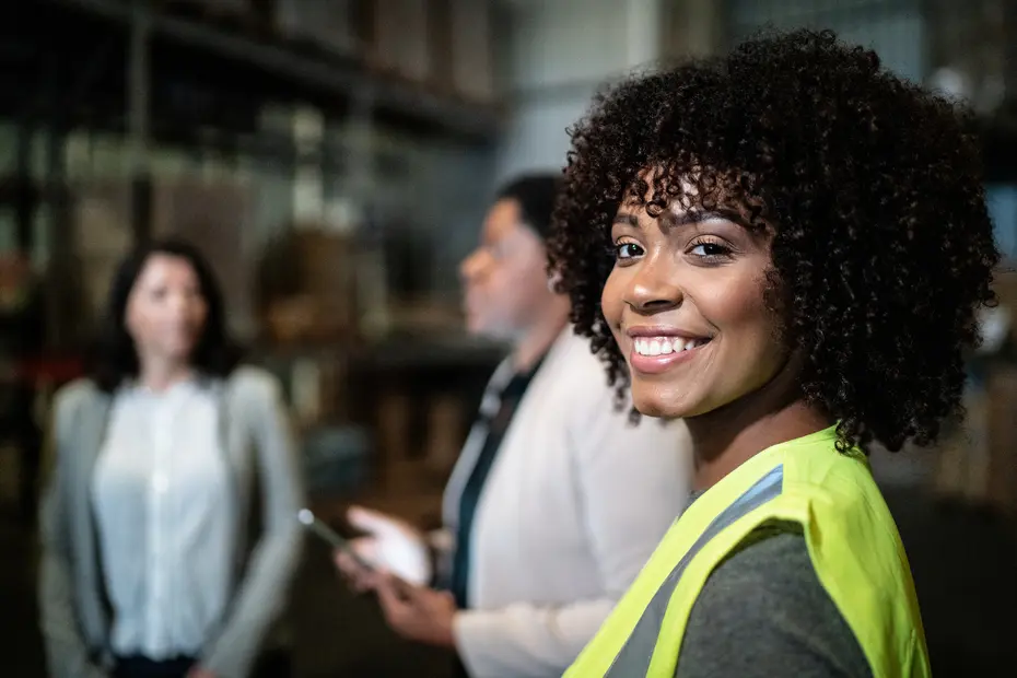 A smiling woman wearing a bright yellow safety vest stands in the foreground, looking at the camera. In the background, two other women are engaged in conversation. The setting appears to be a warehouse or industrial environment.