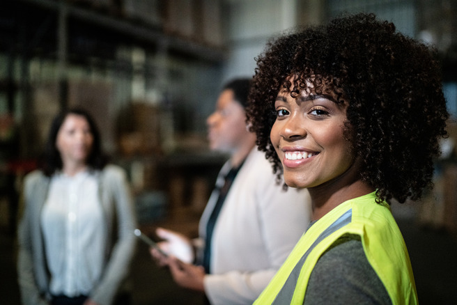 A smiling woman wearing a bright yellow safety vest stands in the foreground, looking at the camera. In the background, two other women are engaged in conversation. The setting appears to be a warehouse or industrial environment.