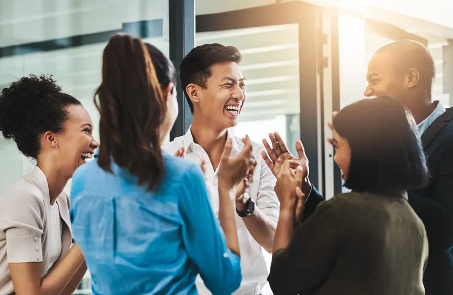 A diverse group of six people are engaged in a joyful conversation, smiling and laughing together. They appear to be in a modern office environment, with sunlight streaming through large windows. The group displays a sense of camaraderie and connection.