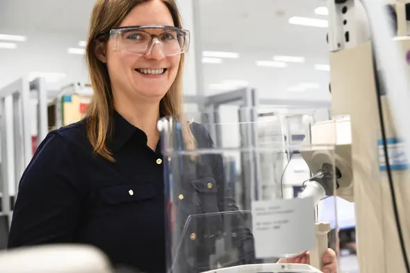 A smiling woman wearing safety glasses stands next to laboratory equipment. She is in a bright, modern lab environment, with shelves and tools visible in the background.