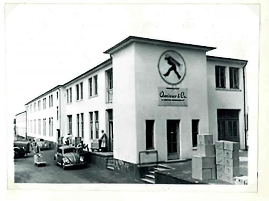A black-and-white photo of a two-story building labeled with a logo featuring a person walking. The structure has multiple windows and a staircase. In front, there are some people and vintage cars, while boxes are stacked nearby.