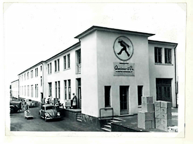 A black-and-white photo of a two-story building labeled with a logo featuring a person walking. The structure has multiple windows and a staircase. In front, there are some people and vintage cars, while boxes are stacked nearby.
