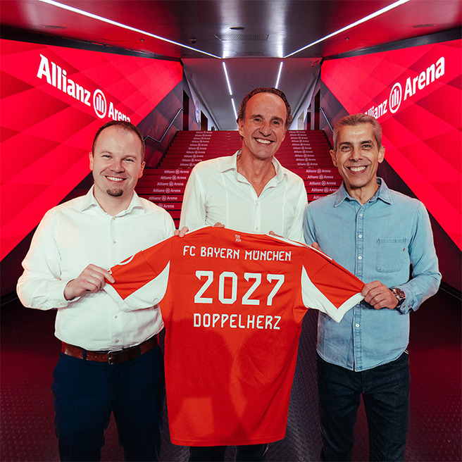 Three men stand together in a hallway at Allianz Arena, holding up a red FC Bayern München jersey featuring the year "2027" and "DOPPELHERZ." The background shows illuminated stairs leading to the arena, emphasizing the team's vibrant branding.