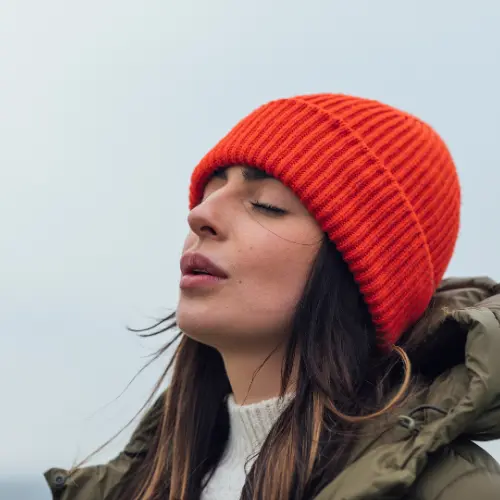 A woman with long, dark hair wears an orange knitted beanie and a green jacket. She closes her eyes, appearing to enjoy a moment of calm outdoors, with a soft expression on her face, suggesting peace or contemplation. The background is neutral and blurred.