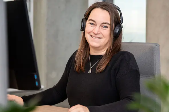 A woman with long brown hair is smiling while wearing headphones at a desk. She is sitting in front of a computer monitor, with a light-colored surface and plants in the foreground. The background features a modern office setting.