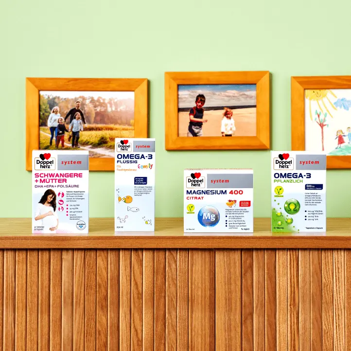 Five health supplement boxes are displayed on a wooden shelf against a green wall. The products include "Schwangere + Mütter," "Magnesium 400 Citrat," "Omega-3 Flushing," and "Omega-3 Pflanzenöl," each with distinct labels highlighting their benefits for health. Framed photos are visible in the background.