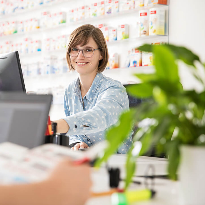 A friendly young woman with short hair and glasses smiles while sitting at a desk in a pharmacy. Behind her is a wall filled with various medication packages. She is reaching out, possibly to assist a customer, with a plant visible in the foreground.