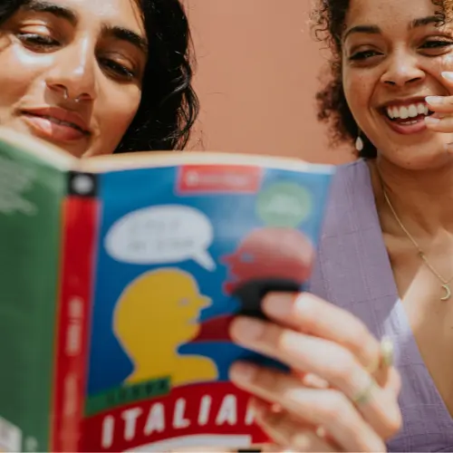 Two women are engaged in reading a colorful book titled "Learn Italian." One woman is smiling while looking at the pages, and the other is focused on the text. The background features a soft, muted color that complements their joyful interaction.