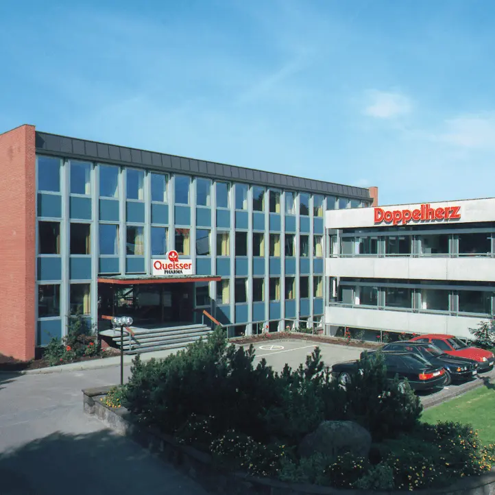 The image shows the exterior of a modern building, divided into two sections with distinct signage. The left side features "Quellesser" in a prominent display, while the right side has "Doppelherz." There are several cars parked in front and greenery surrounding the entrance.