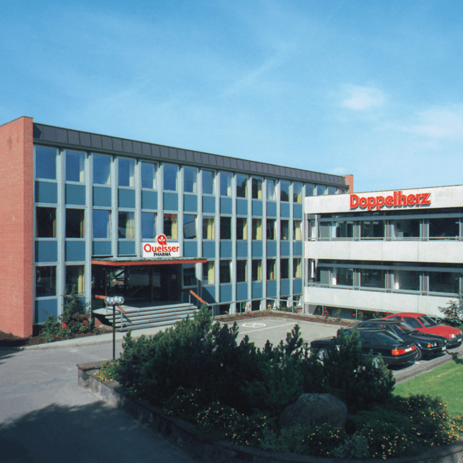 The image shows the exterior of a modern building, divided into two sections with distinct signage. The left side features "Quellesser" in a prominent display, while the right side has "Doppelherz." There are several cars parked in front and greenery surrounding the entrance.