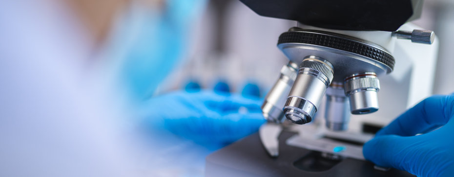 A close-up of a laboratory microscope with metallic lenses, being adjusted by a person wearing blue gloves. The focus is on the microscope, suggesting a scientific examination or research setting.