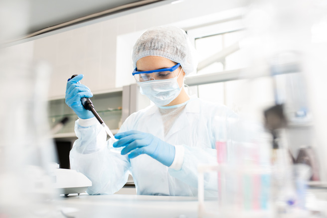 A scientist in a laboratory wearing a lab coat, gloves, protective goggles, and a face mask is using a pipette to transfer a liquid in a lab setting. Test tubes and laboratory equipment are visible in the background.