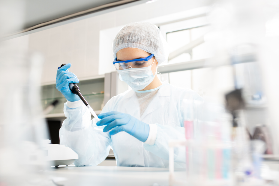 A scientist in a laboratory wearing a lab coat, gloves, protective goggles, and a face mask is using a pipette to transfer a liquid in a lab setting. Test tubes and laboratory equipment are visible in the background.