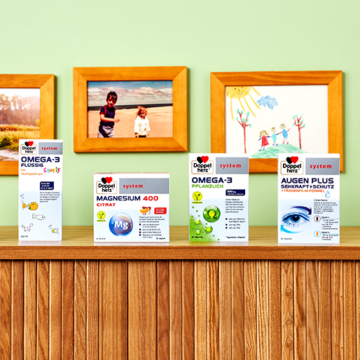 A wooden shelf displays various health supplement boxes labeled "Omega-3 Flüssig," "Magnesium 400 Citrat," "Omega-3 Pflanzenöl," and "Augen Plus." In the background, colorful children's drawings and two framed photos are visible against a green wall.