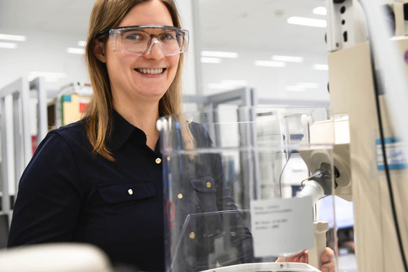 A smiling woman wearing safety glasses stands next to laboratory equipment. She is in a bright, modern lab environment, with shelves and tools visible in the background.