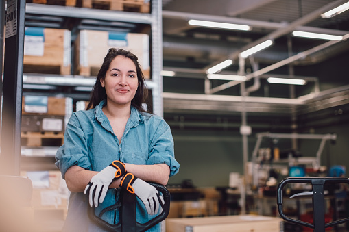 A woman wearing a light blue shirt and gloves stands confidently in a warehouse setting, leaning on a pallet jack. The background features shelves of boxes and various industrial equipment, indicating a work environment.